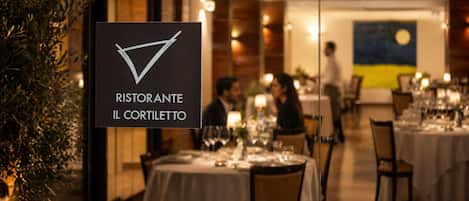 L’intérieur d’un restaurant, avec des tables dressées pour le service, une enseigne indiquant le nom « RISTORANTE IL CORTILETTO », et une personne à l’arrière-plan.