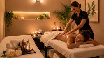 A spa room with two massage tables, a wall-mounted shelf with plants, candles, and spa products.