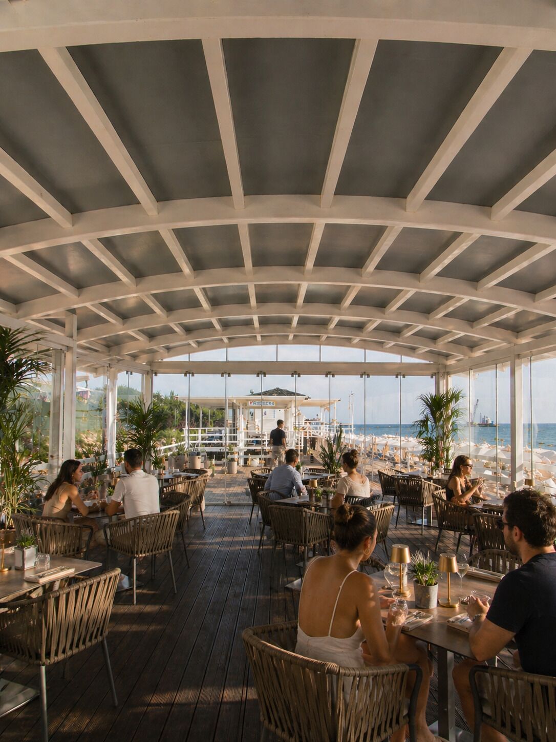 An outdoor dining area with tables and chairs, a covered ceiling, and a view of the sea.