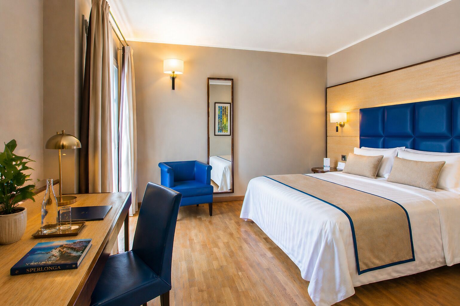 A hotel room with a bed, a desk, a chair, a lamp, and a potted plant.