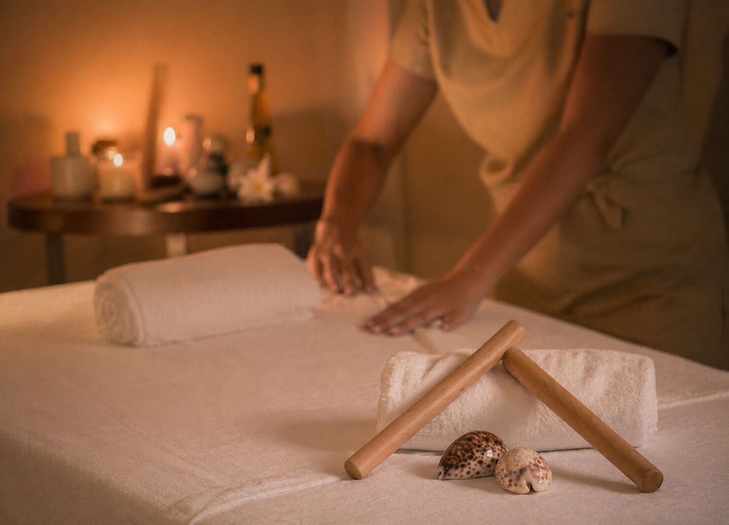 Une personne est en train de préparer une table de massage avec des serviettes et des bâtonnets en bois, dans une pièce à l’atmosphère chaleureuse, éclairée par des bougies.