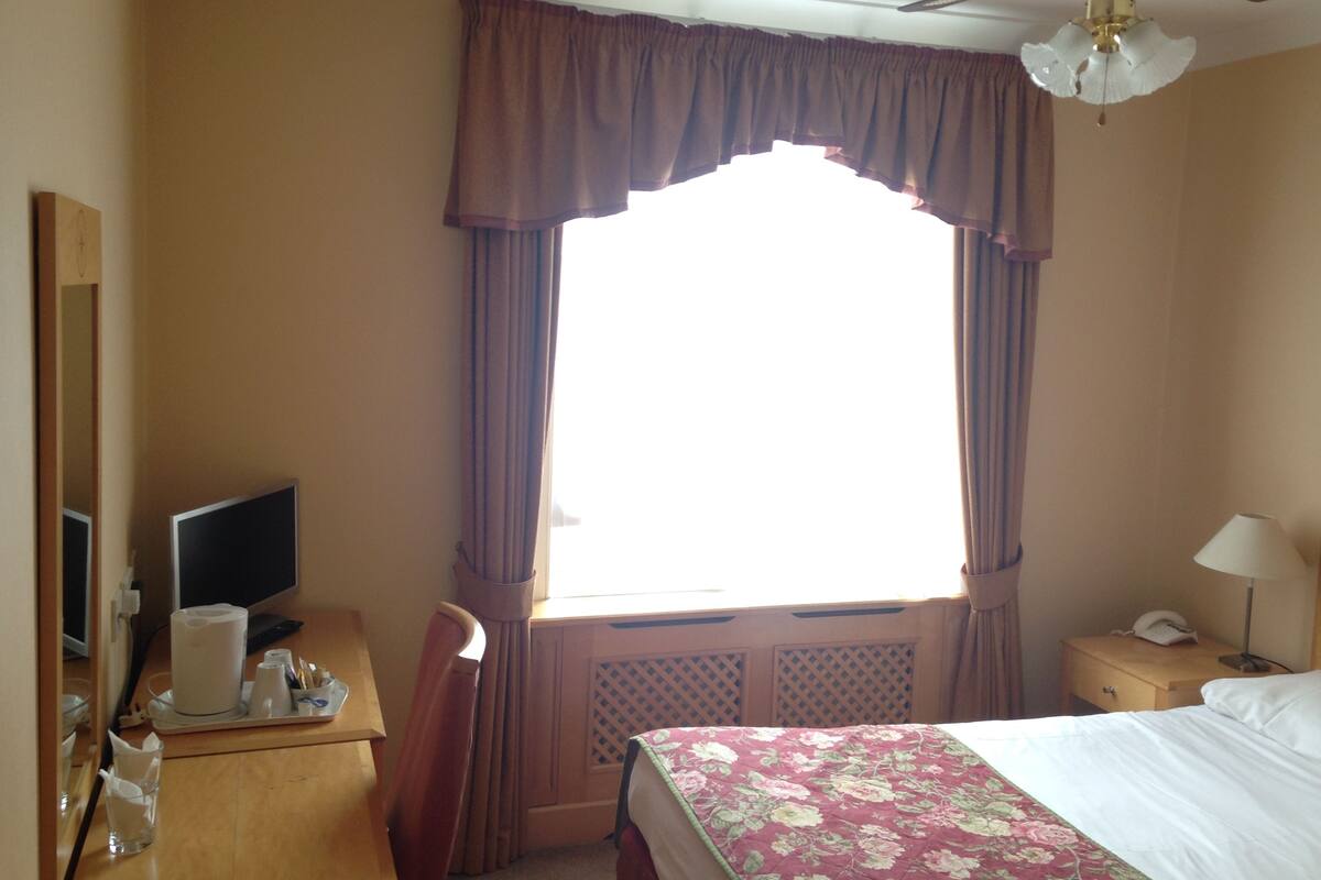 A hotel room with a bed, a desk with a computer, a lamp, and a window with curtains.