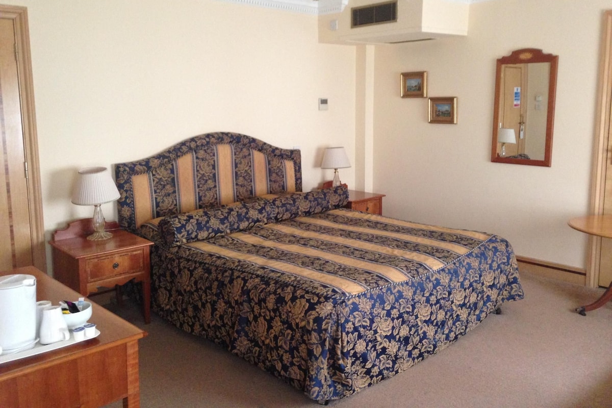 A bedroom with a large bed, two bedside tables, a wooden dresser, a mirror, and a small table with a lamp.