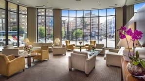 Un vestíbulo de hotel con amplios ventanales, varias zonas de estar y una mesa central con un jarrón de flores.
