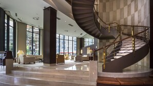Un vestíbulo amplio con una escalera curva, ventanas grandes y un área de descanso con sofás y lámparas.