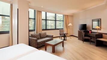 Una habitación de hotel con una cama grande, un sofá, una mesita de centro, un escritorio con una silla, un televisor y ventanas amplias con cortinas.