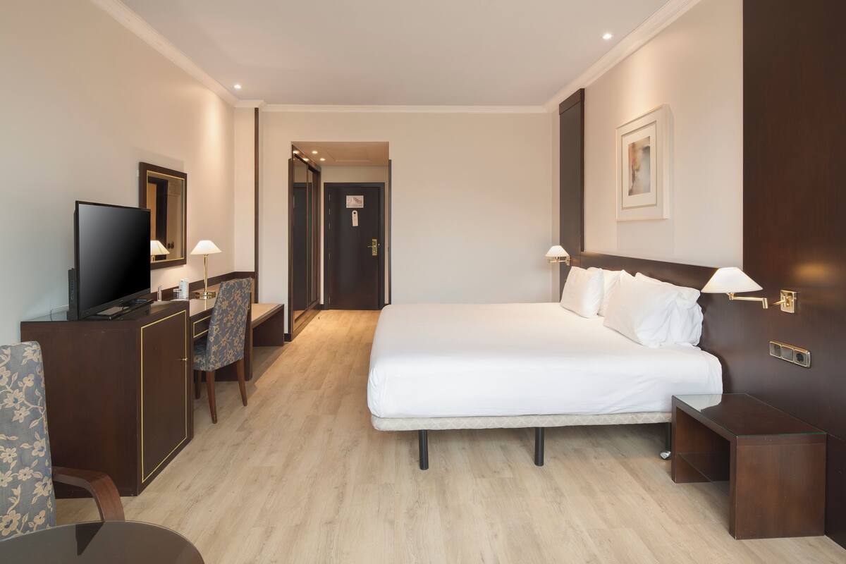 A hotel room with a bed, bedside tables, a desk with a television, a chair, and a mirror.