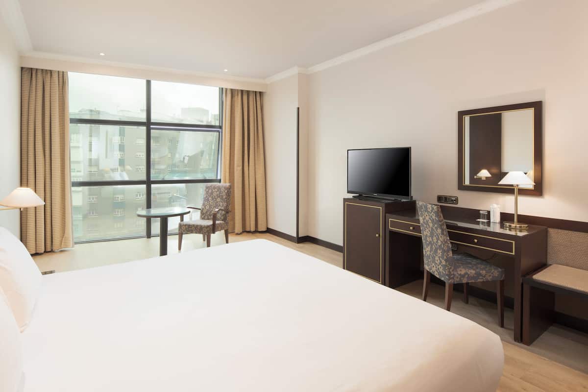 A hotel room with a large bed, a desk with a television, a chair, a small table, and a large window with curtains.