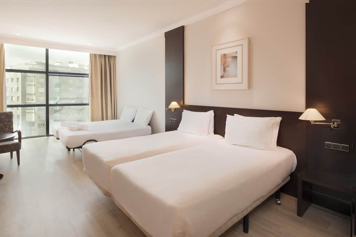 A hotel room with two beds, a desk, a chair, a large window, and a framed picture on the wall.