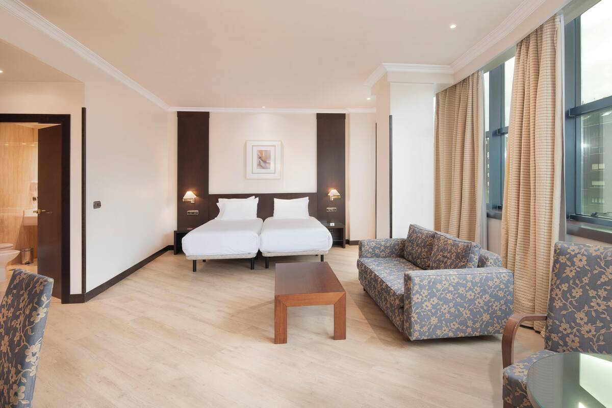 A hotel room with a bed, two bedside tables, a sofa, a coffee table, and a view of the city through large windows.