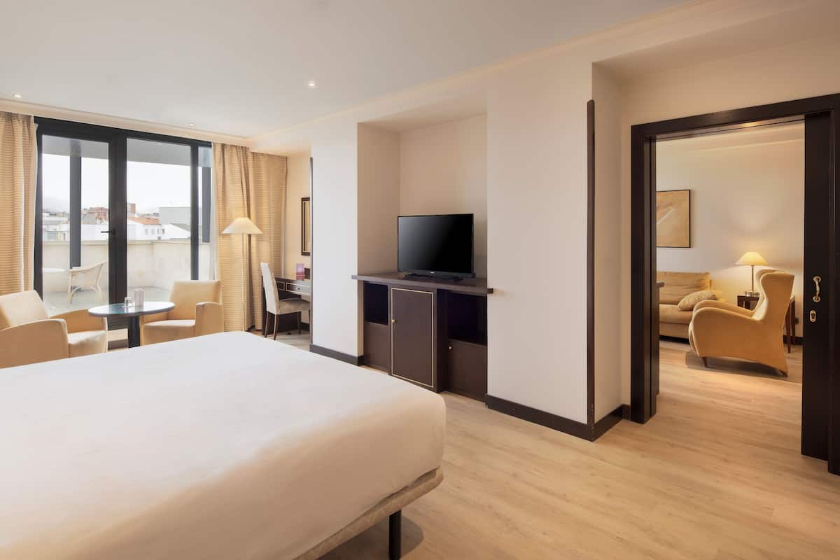 A hotel room with a bed, a desk with a television, a chair, and a view of the city through large windows.