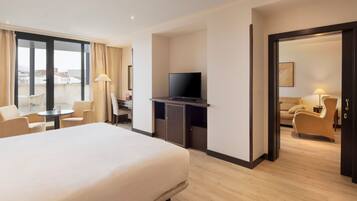 A hotel room with a bed, a desk with a television, a chair, and a view of the city through large windows.