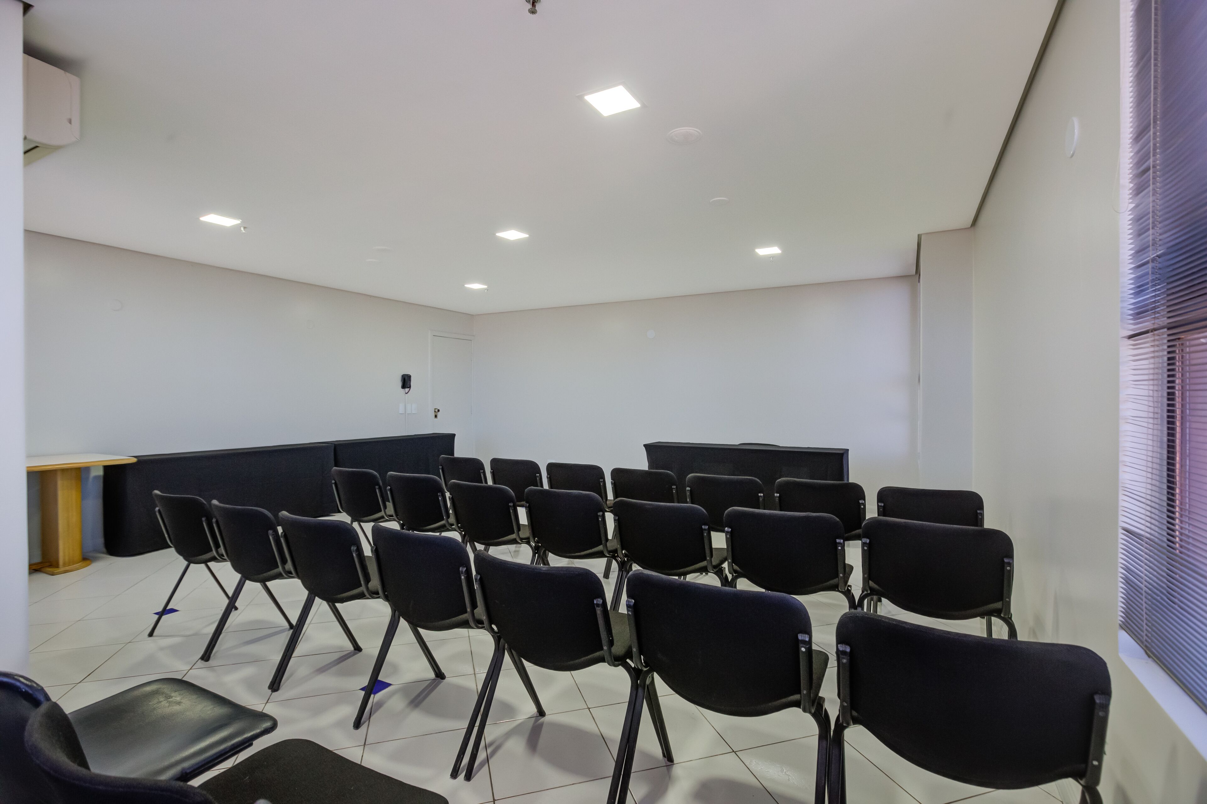 Un salón de conferencias con filas de sillas negras, un atril y un amplio ventanal con persianas.