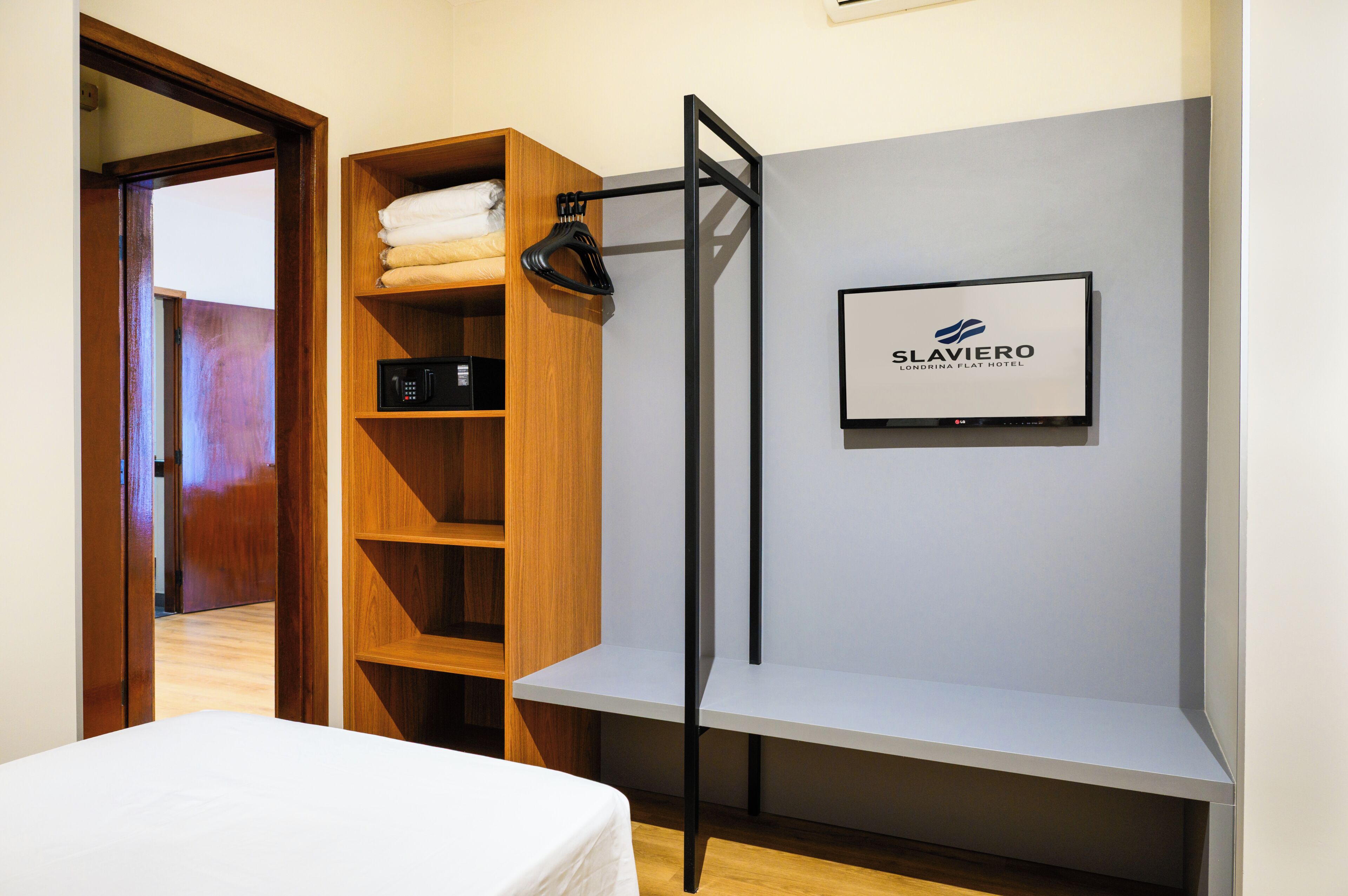 A hotel room with a wooden wardrobe, a bed, a wall-mounted TV, and a door leading to another room.