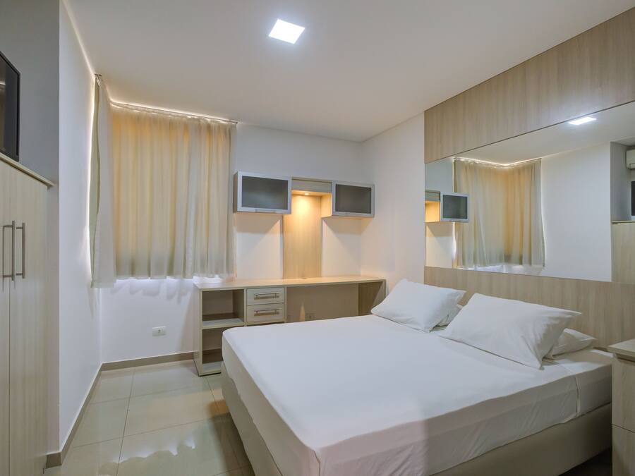 A modern hotel room with a bed, a desk, a chair, a flat-screen TV, a mirror, and a wardrobe.