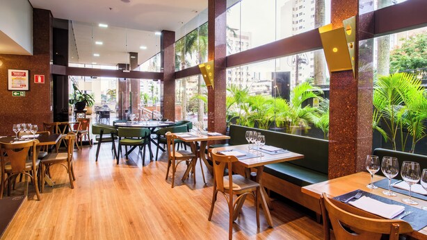 A restaurant with wooden tables and chairs, large windows with a view of palm trees and buildings, and a modern interior design. - SLAVIERO Londrina (Londrina)