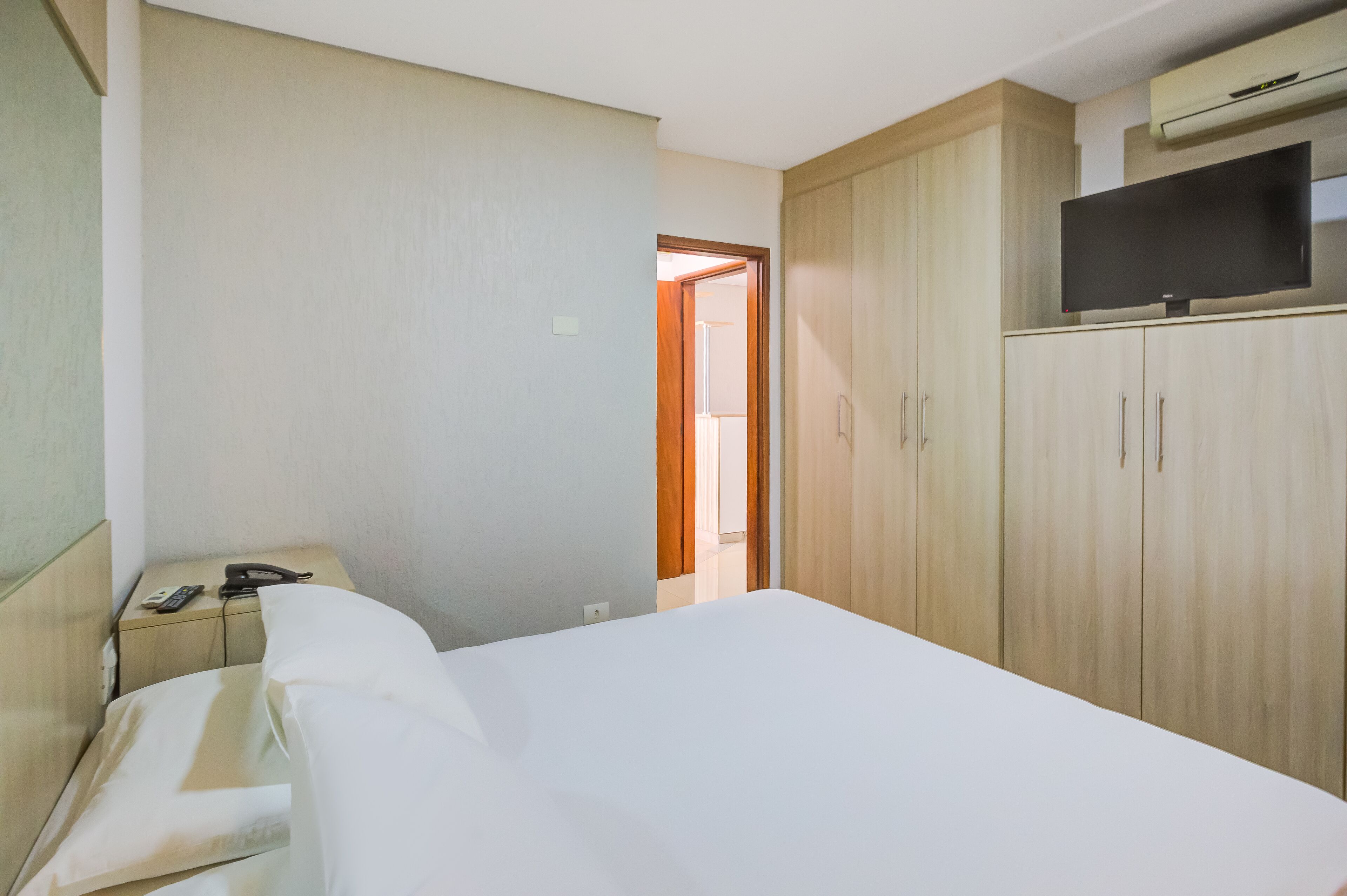 A hotel room with a bed, a desk with a telephone and a remote control, a wardrobe, and a television on top of the wardrobe.