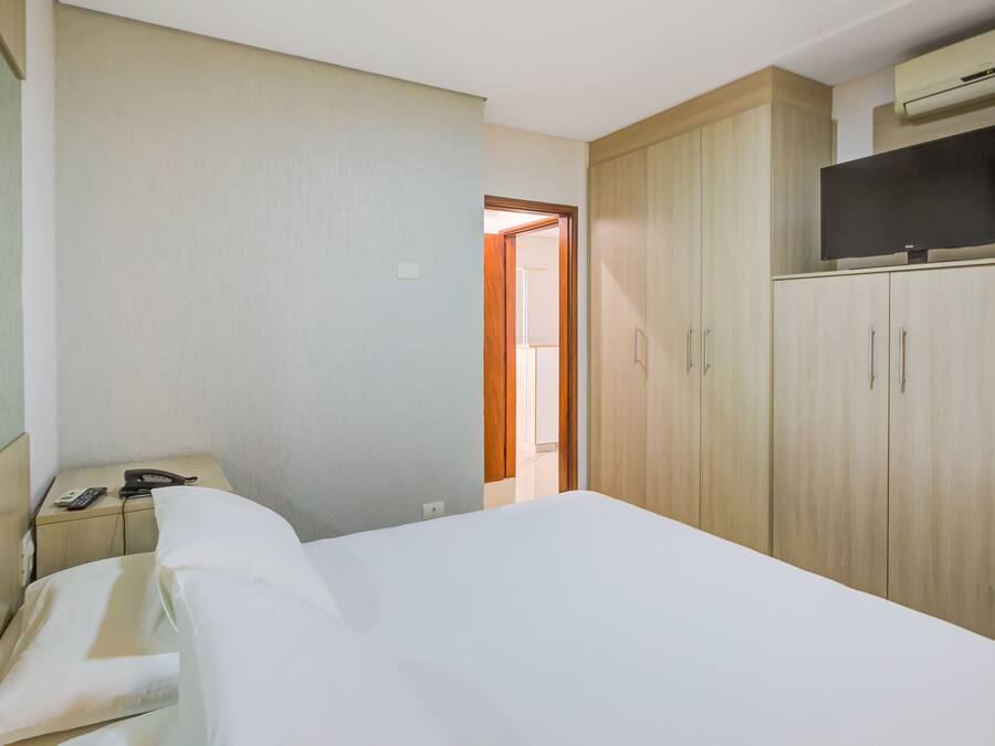 A hotel room with a bed, a desk with a telephone and a remote control, a wardrobe, and a television on top of the wardrobe.