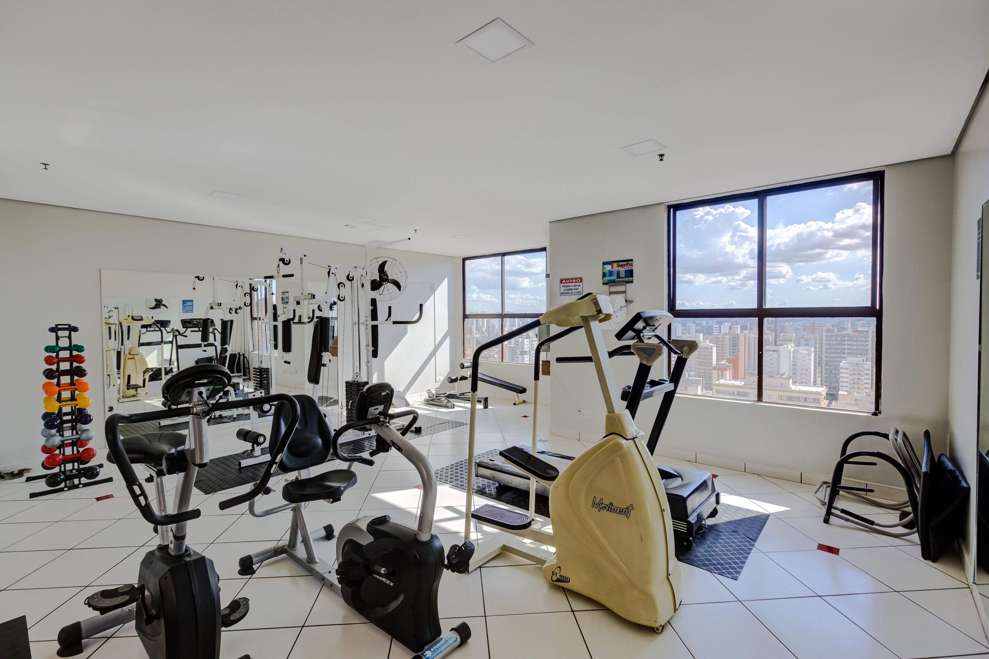 Un gimnasio con diversos equipos de ejercicio, amplias ventanas con vista a la ciudad y un ventilador de techo.