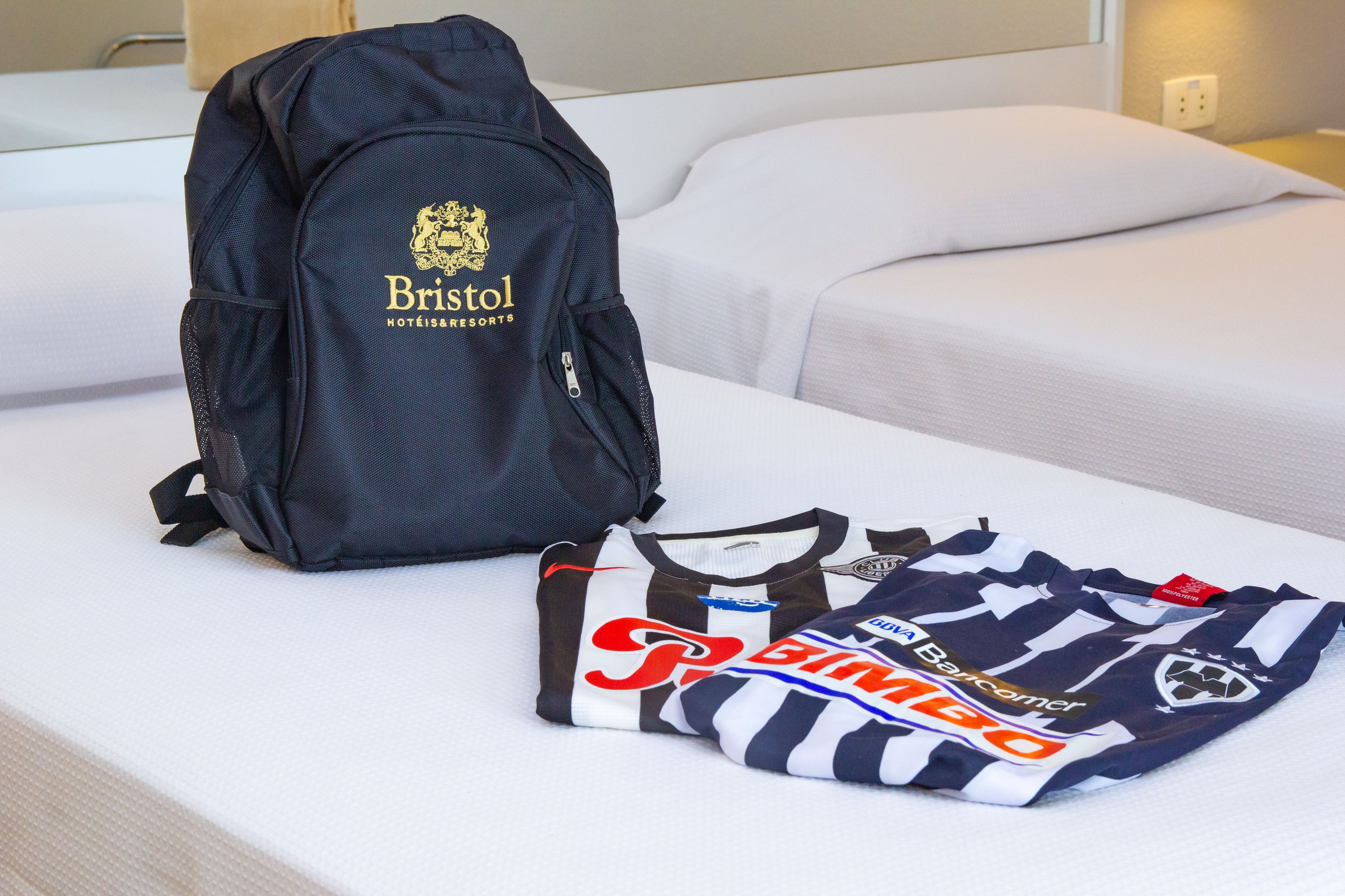 A neatly made bed with a white comforter and a black backpack with the logo "Bristol Hotels & Resorts" on it.