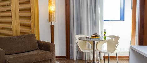 A room with a brown sofa, a table with two white chairs, a lamp, and a window with curtains.