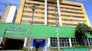 Un edificio alto con fachada azul y amarilla, una entrada de ladrillo verde con la leyenda 'Bristol Brasil 500 Business & Hotel', y un cartel de 'CAIXA' en la entrada.