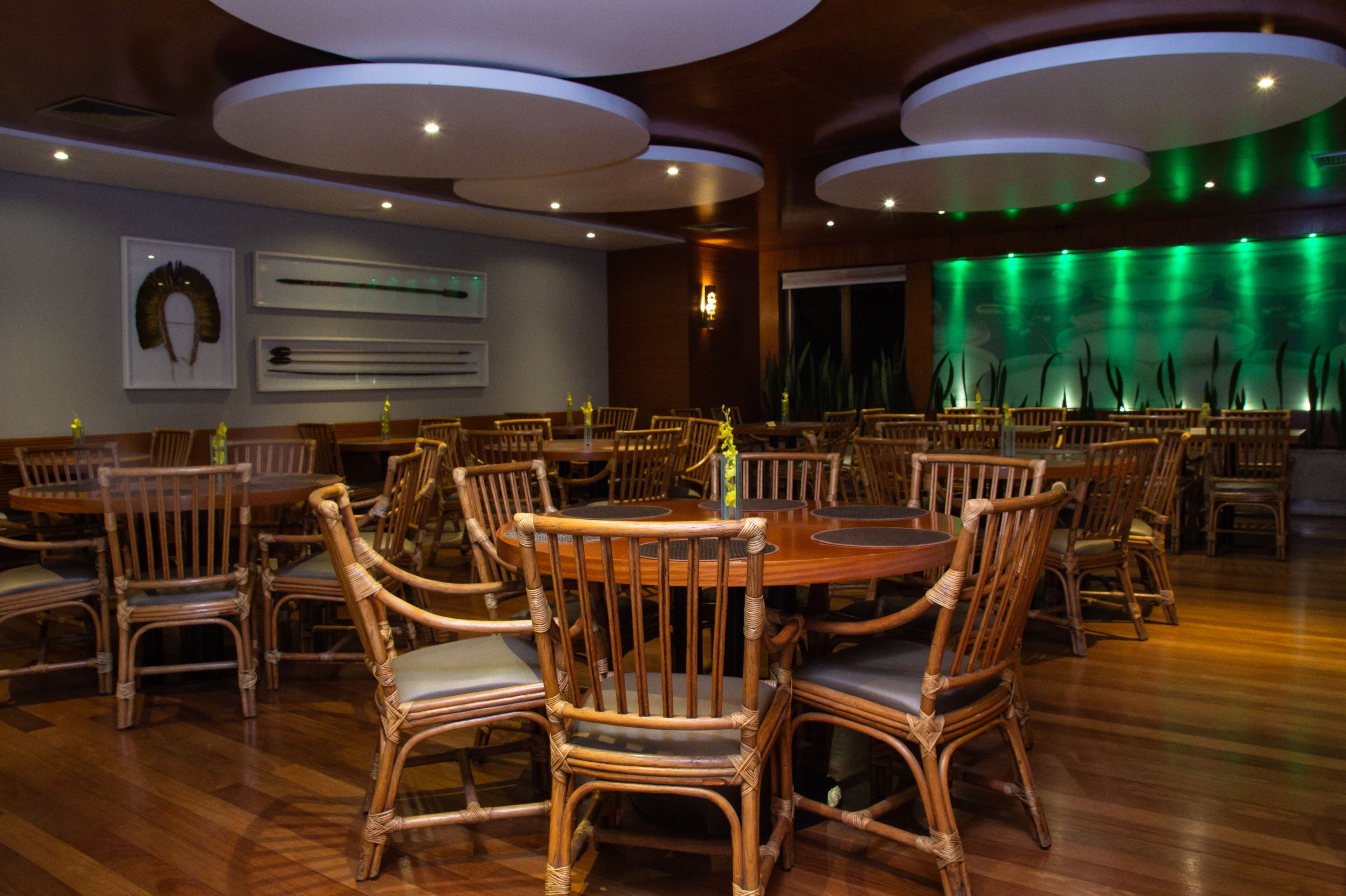 A restaurant with round tables and wicker chairs, featuring a modern ceiling design and a green-lit wall.