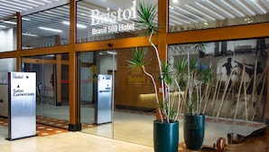 La entrada del Hotel Bristol Brasil 500, con un cartel del hotel y áreas comerciales, plantas grandes en macetas y un banco de madera.