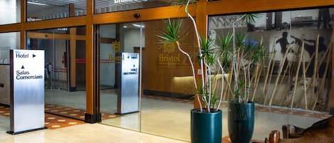 The entrance of the Bristol Brasil 500 Hotel with a sign for the hotel and commercial areas, large potted plants, and a wooden bench.