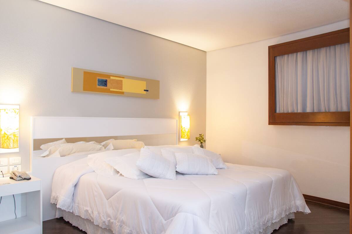 Quarto de hotel com uma cama grande, mesa de cabeceira com telefone e luminária fixa na parede.