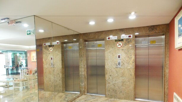 A modern elevator area with marble flooring and glass walls, featuring four elevators and a visible emergency exit sign. - Duomo Park Hotel (Curitiba)
