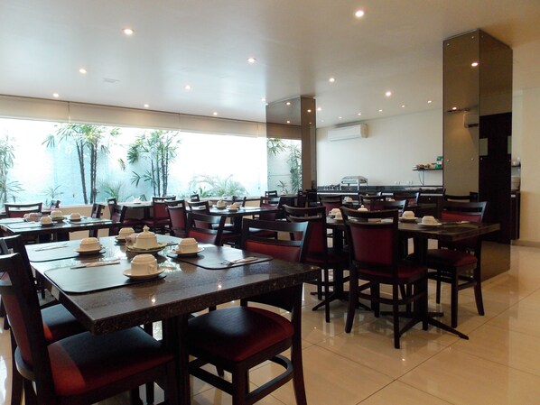Un restaurante con varias mesas y sillas, una zona de cocina y amplios ventanales con vistas a palmeras.
