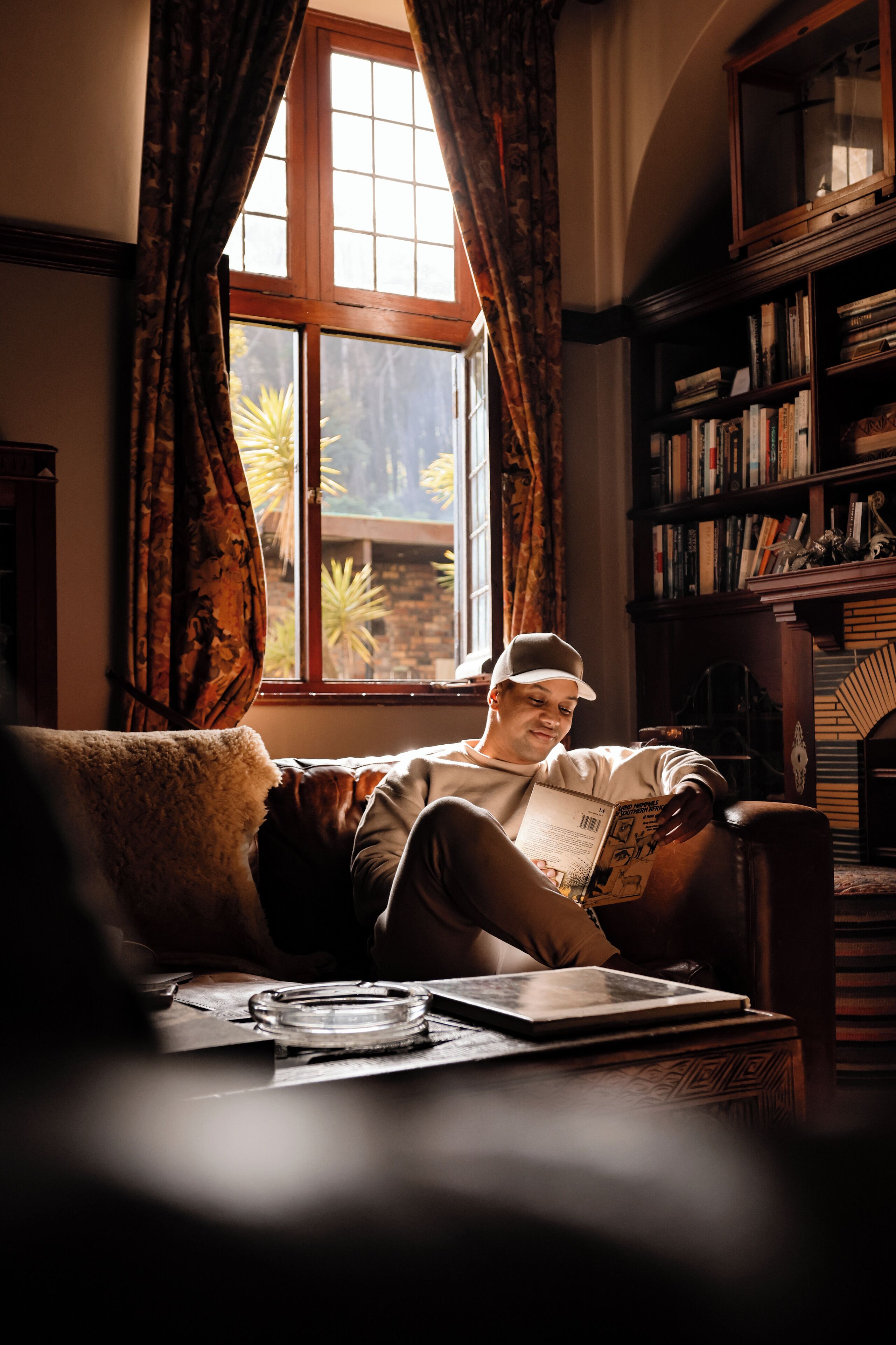A person reading a book in a room with a large window, a bookshelf, and a patterned rug.