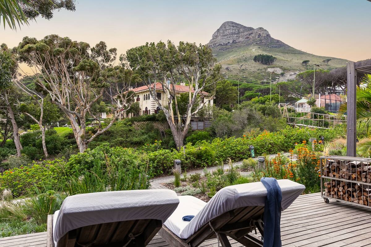 A wooden deck with two lounge chairs, a garden with various plants, and a view of a mountain and a house in the background.