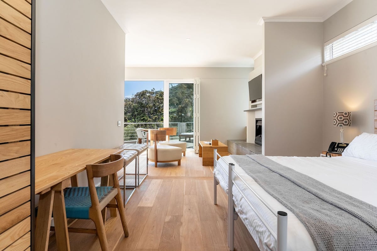 A modern hotel room with a wooden desk, a bed, a chair, and a balcony with a view of trees.