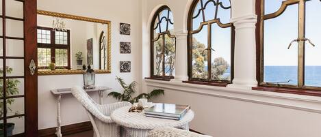 Una habitación con muebles de mimbre blanco, un espejo grande y ventanas arqueadas con vista al mar.