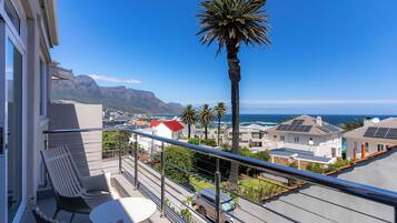 A balcony with a view of a coastal town, a palm tree, and a car parked on the street.