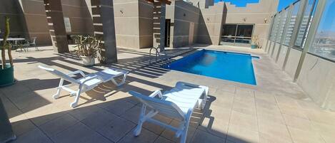 Uma área de piscina no terraço com cadeiras de lounge brancas, piscina e uma estrutura de pérgola.