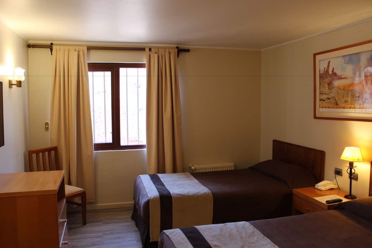 A hotel room with two beds, a wooden desk, a chair, a window with curtains, a radiator, and a painting on the wall.