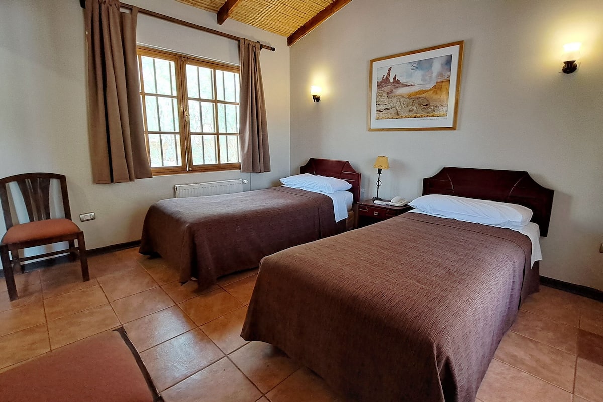 A hotel room with two beds, a chair, a painting on the wall, and a window with curtains.