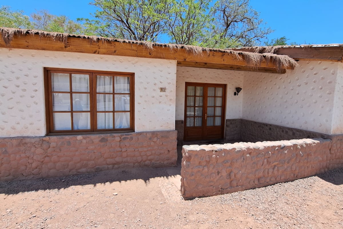 A traditional adobe house with a thatched roof, a stone foundation, and a window with a wooden frame and curtains.
