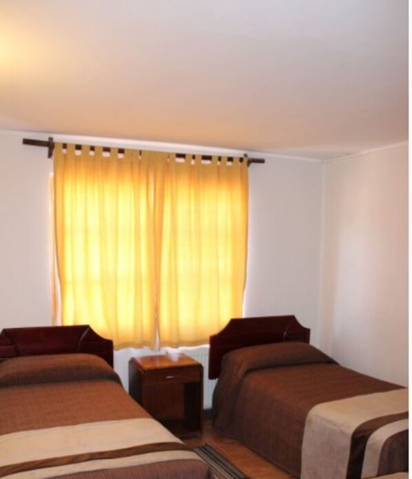 A hotel room with two single beds, a small bedside table, and a window with yellow curtains.