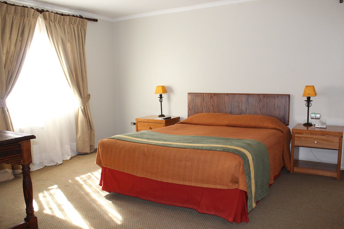 A hotel room with a large bed, two bedside tables with lamps, a wooden nightstand, and a window with curtains.