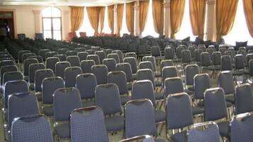 A large hall with rows of blue chairs, a high ceiling, and large windows with curtains.