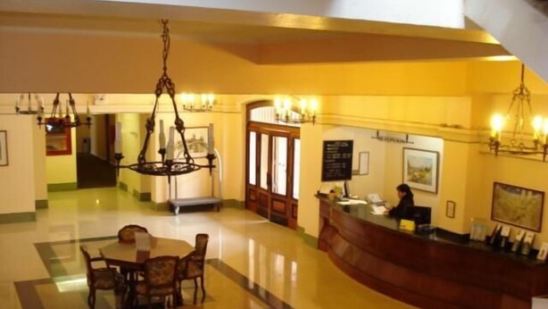 Vestíbulo de hotel con una mesa de recepción, un candelabro, un área de comedor con sillas y una escalera.