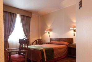 Una habitación de hotel con cama, dos mesitas de noche, una silla y un ventanal con cortinas.