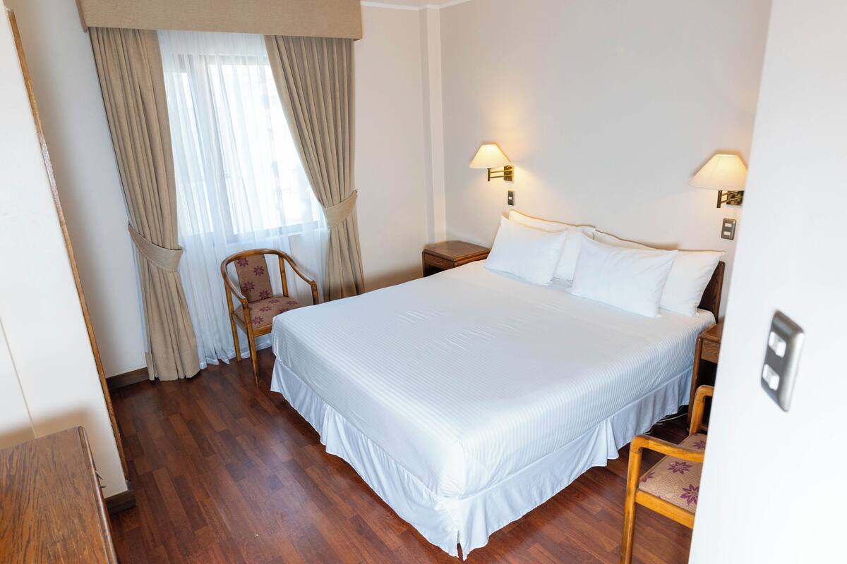 Habitación de hotel con una cama grande, suelo de madera, una silla y dos lámparas fijadas a la pared.