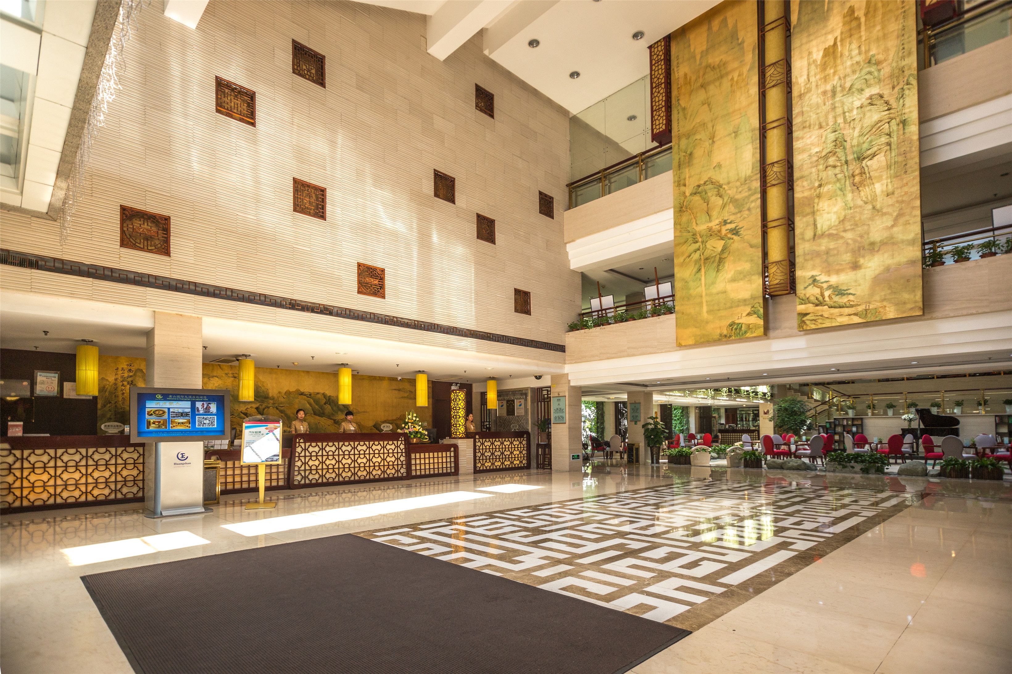 A spacious lobby with a high ceiling, a reception desk, a digital information board, and a patterned floor.