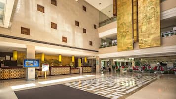 A spacious lobby with a high ceiling, a reception desk, a digital information board, and a patterned floor.