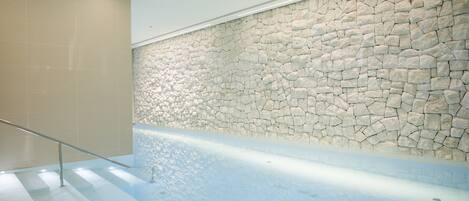 Un área de piscina interior moderna con pared de piedra y barandilla de vidrio.